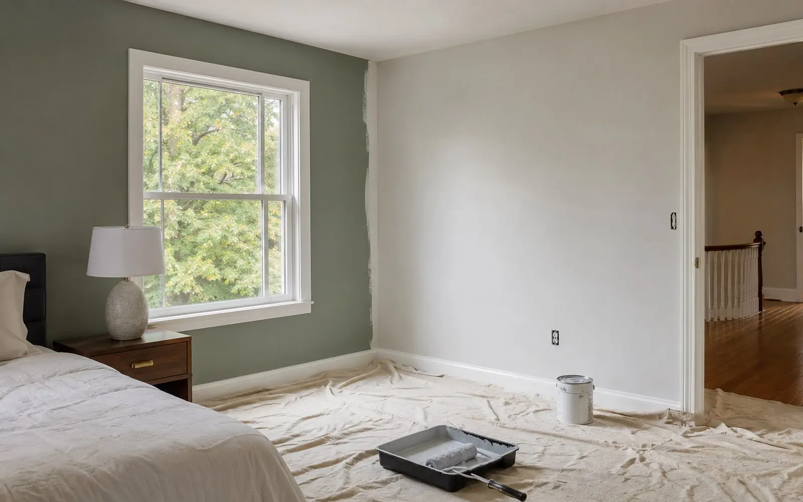 A partly redecorated bedroom with fresh paint on two walls, a drop cloth over the floor, a roller in a tray, and an electrical outlet cover removed.