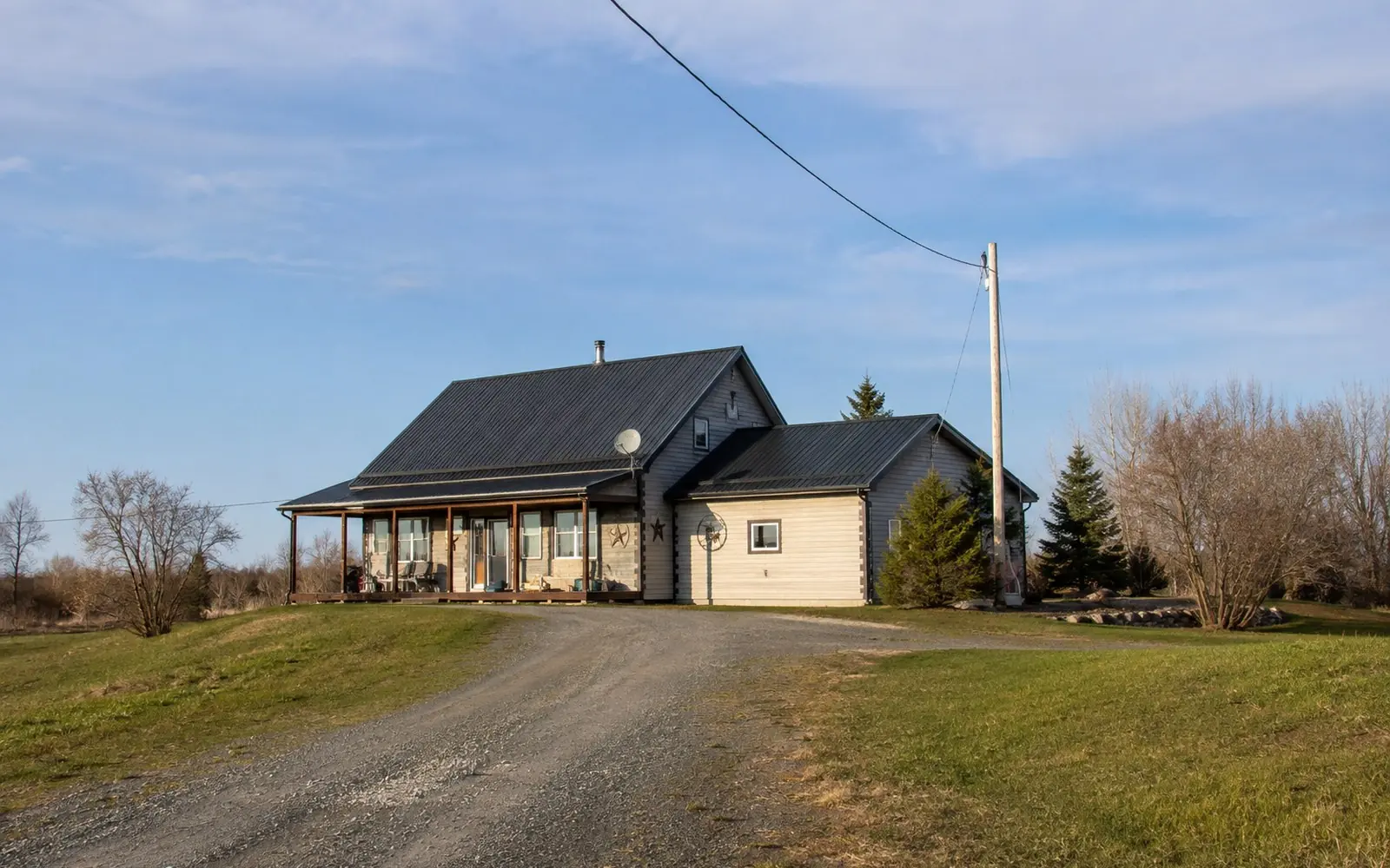 A farmhouse at the end of a gravel road with a satellite dish on the roof and a utility pole carrying a single line to the property.