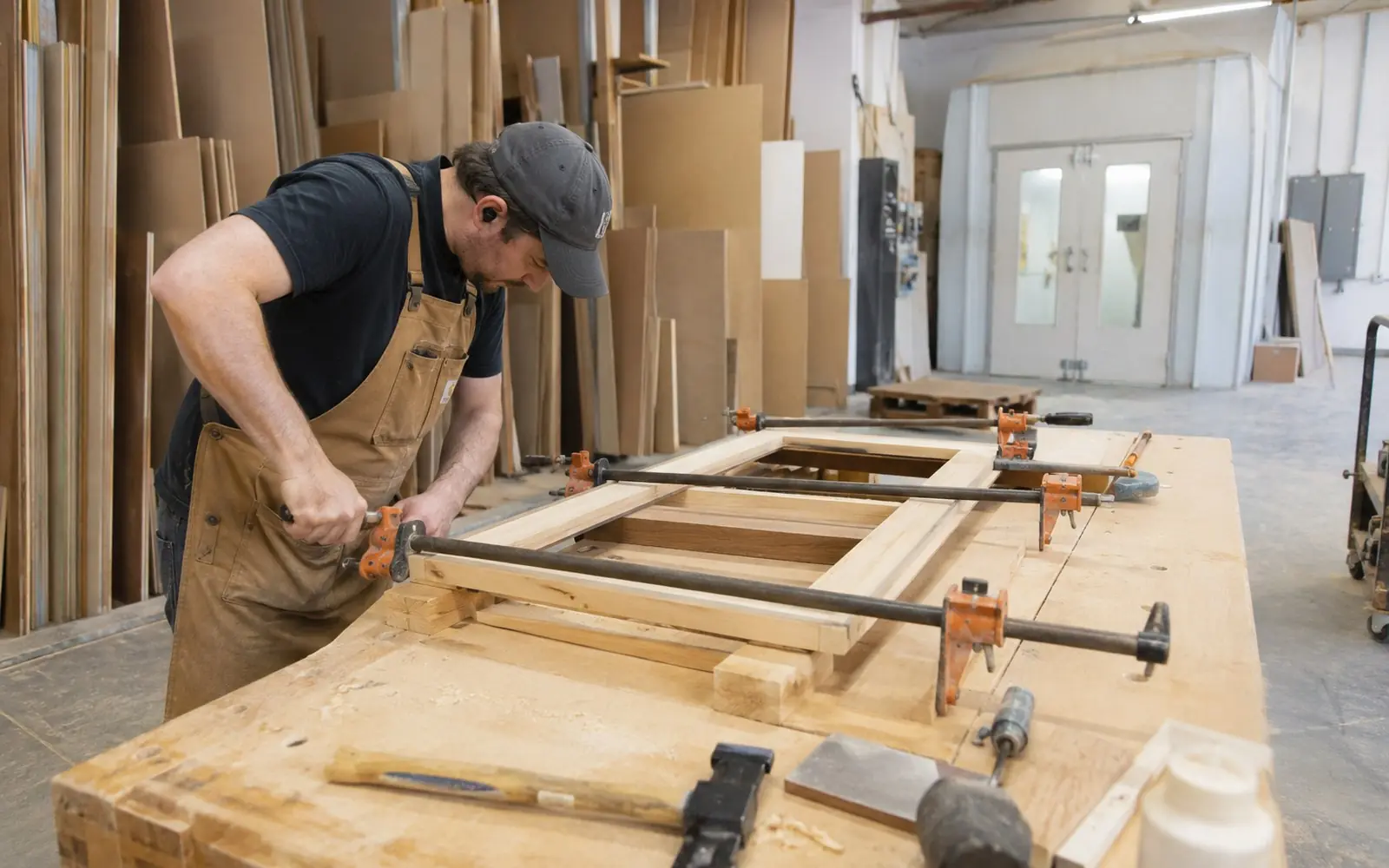 A cabinetmaker in a workshop clamping a face frame at a bench, with sheet goods racked behind and a spray booth visible at the back.