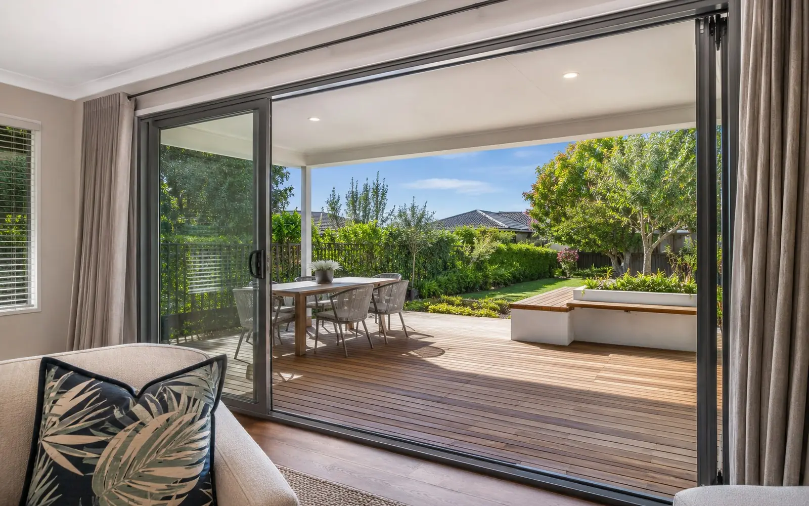 A wide aluminium-framed sliding glass patio door open onto a timber deck, seen from inside a living room in daylight.
