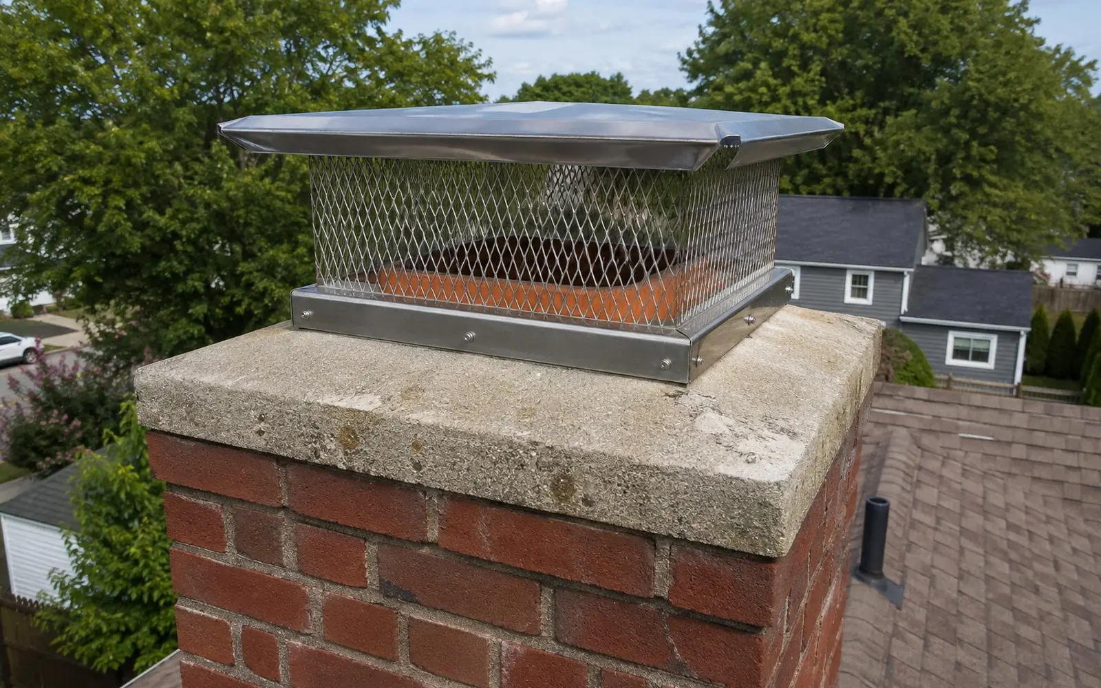 A stainless steel chimney cap with mesh sides fitted over a clay flue tile on top of a brick chimney, photographed from roof level.