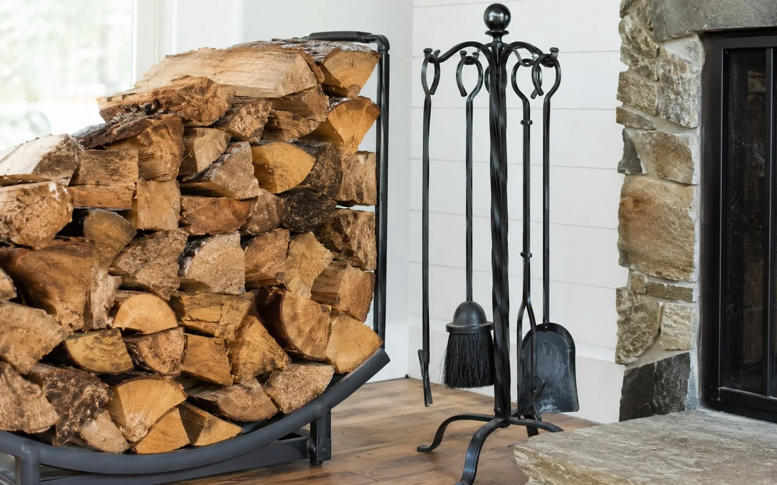 A stack of split firewood beside a hearth, with a black iron tool set of poker, brush, shovel and tongs standing on the floor next to it.