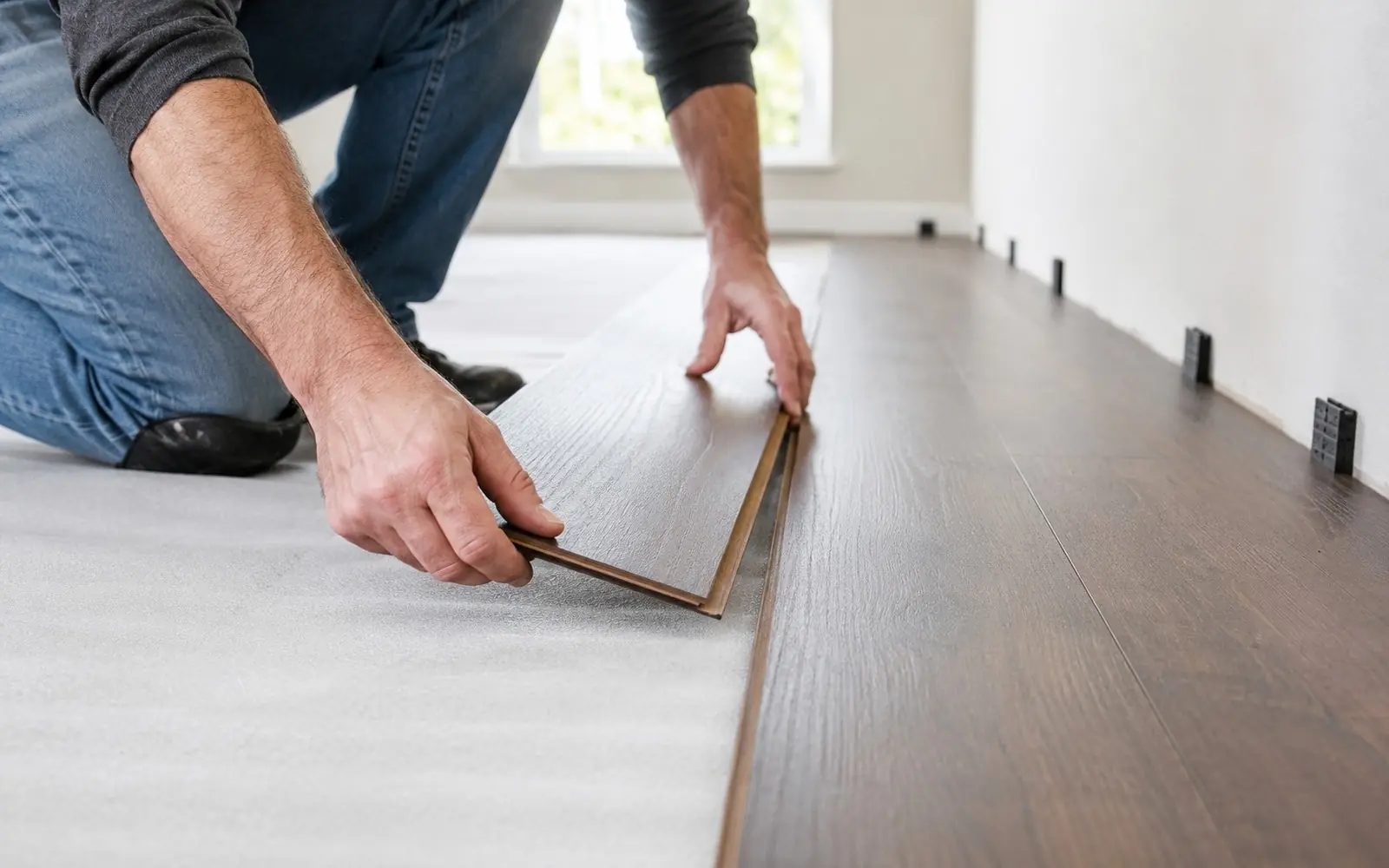 Hands clicking laminate planks over underlayment, spacers at the wall