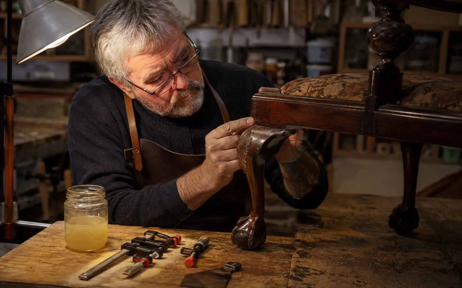 A restorer examining the carved leg of a dark mahogany antique chair under a work lamp, with hide glue, small clamps and a scraper on the bench nearby.