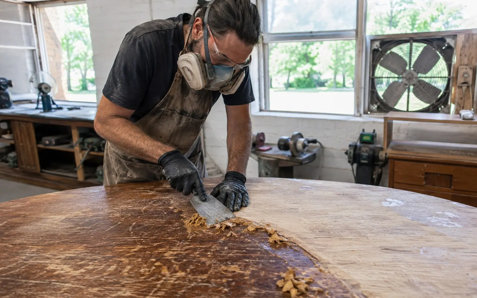 A refinisher in a respirator and gloves scraping softened finish from an old wooden tabletop with a broad blade, in a ventilated shop with the extractor running.