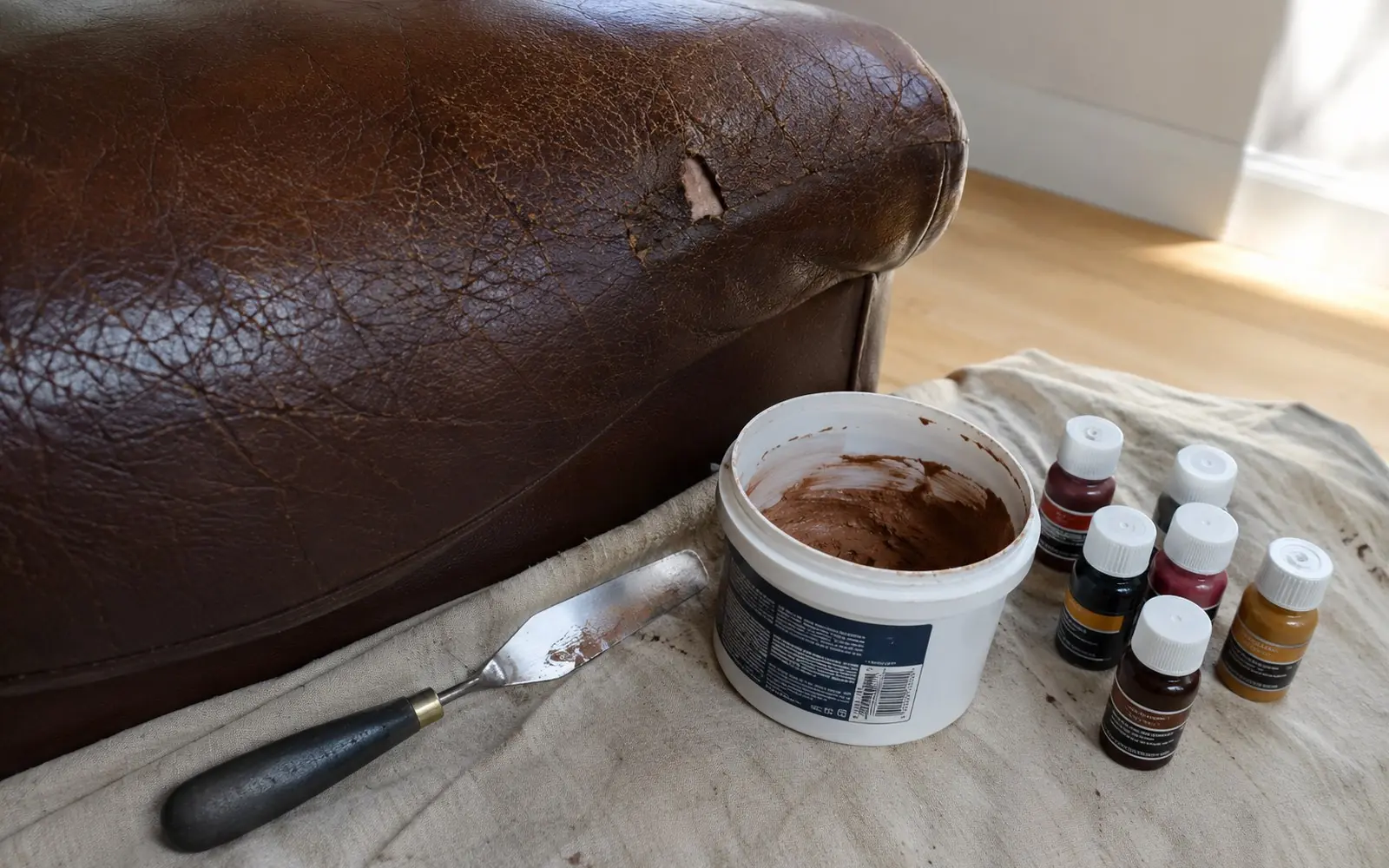 A close-up of a cracked brown leather sofa arm with a small tear, beside a palette knife, a tub of filler compound and colour bottles laid out on a cloth.
