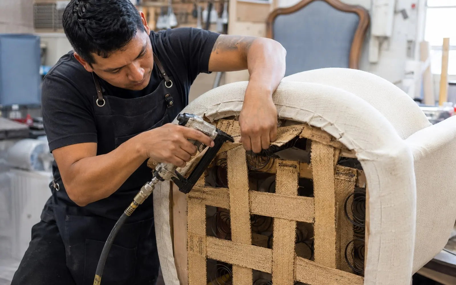 An upholsterer stretching fabric over a chair seat frame with a pneumatic staple gun, with exposed jute webbing and coil springs visible underneath.