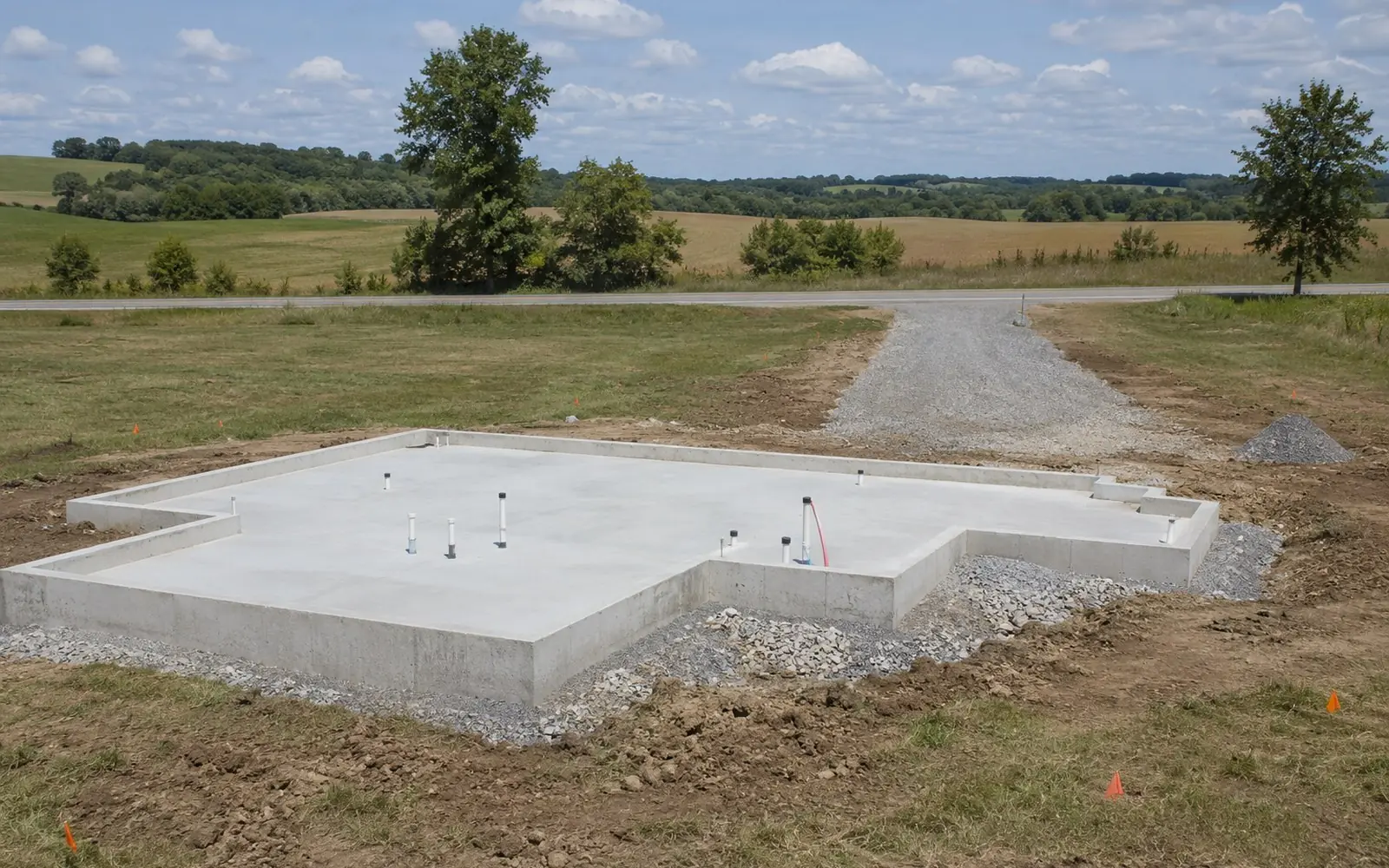 A cleared rural building site with a freshly poured foundation, a gravel construction drive running back to a country road, and orange utility marking flags in the surrounding grass.
