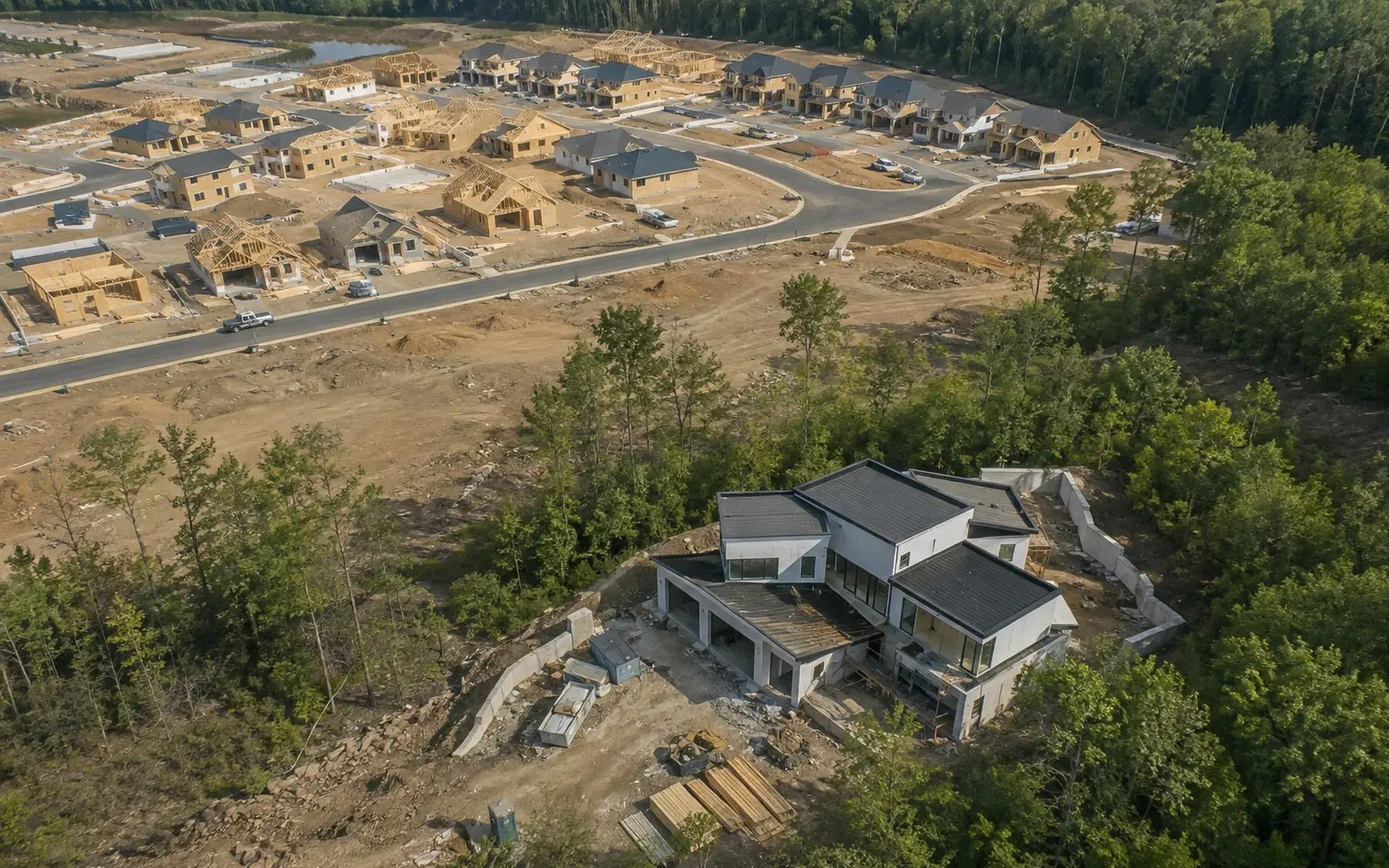 Split view of a new-home subdivision with several near-identical houses at different stages of framing on one side, and a single architect-designed house going up alone on a wooded rural lot on the other.