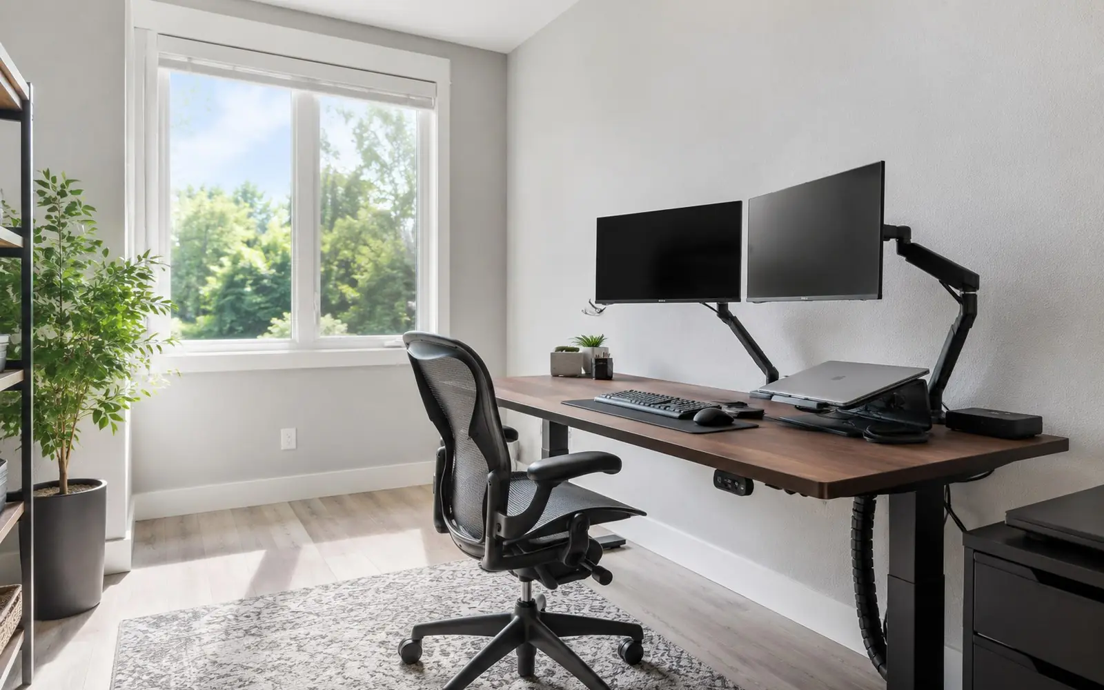 A home office workstation with a mesh task chair pulled up to a height-adjustable desk holding two monitors on arms, a keyboard and a laptop dock.