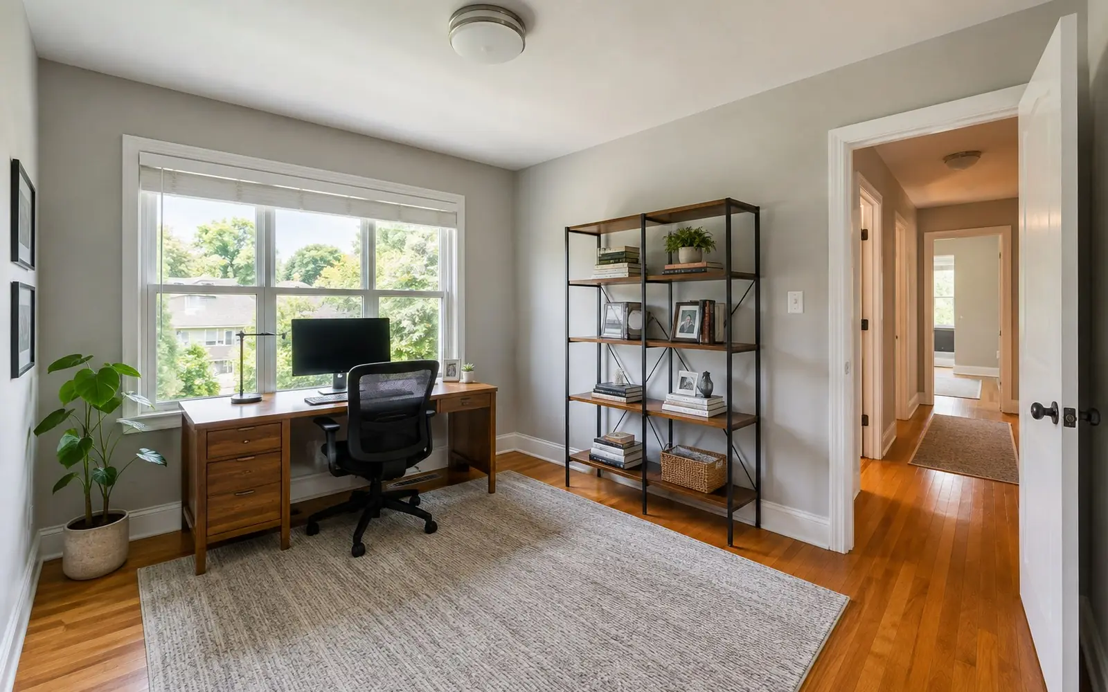 A spare bedroom converted into a home office, with a desk set against a window wall, a tall open shelf unit on the adjacent wall, and a doorway to a hallway.