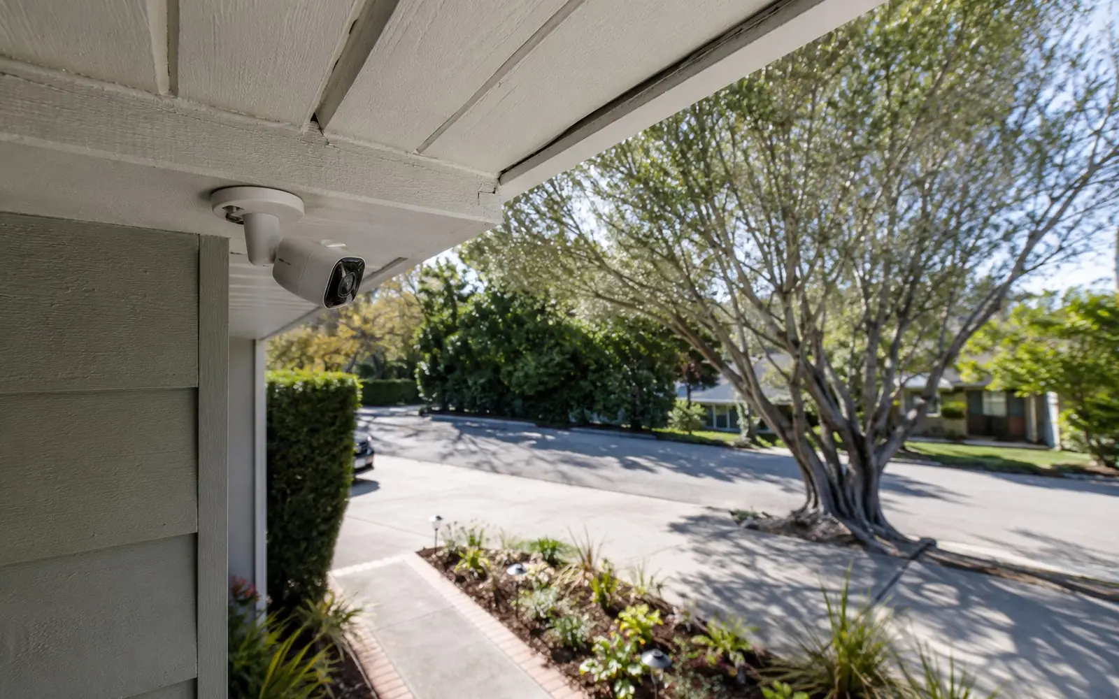 A small white outdoor security camera mounted under a house eave, angled down toward a driveway and front path.