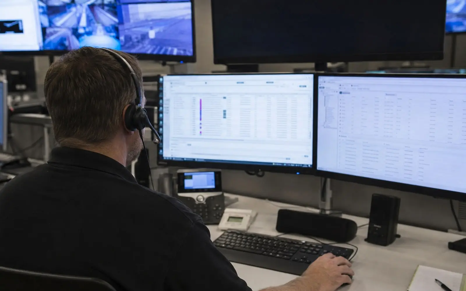 A monitoring station operator wearing a headset at a desk with several screens showing alarm account records.