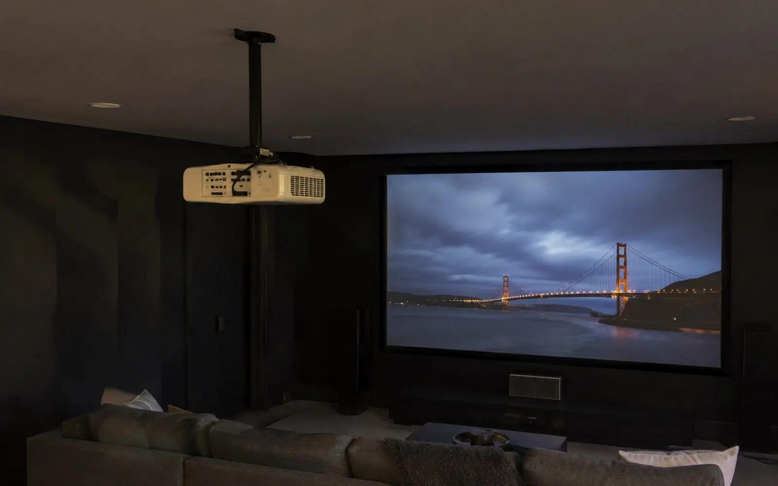 A ceiling mounted projector on a black drop pole throwing an image onto a wide fixed frame screen in a darkened room.