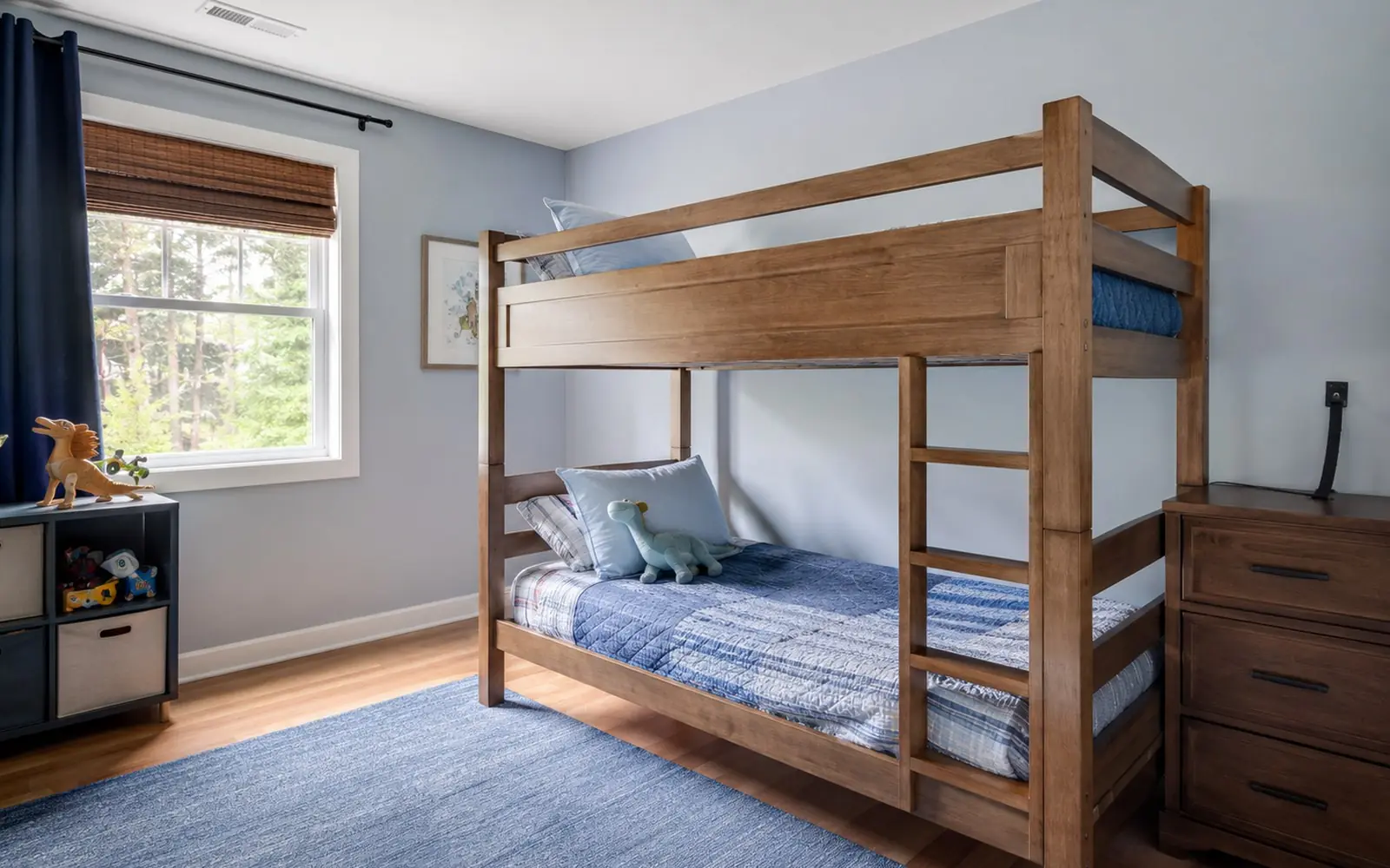 A child's bedroom with a bunk bed showing a full-length guardrail on the side opposite the ladder, and a low dresser strapped to the wall behind it.