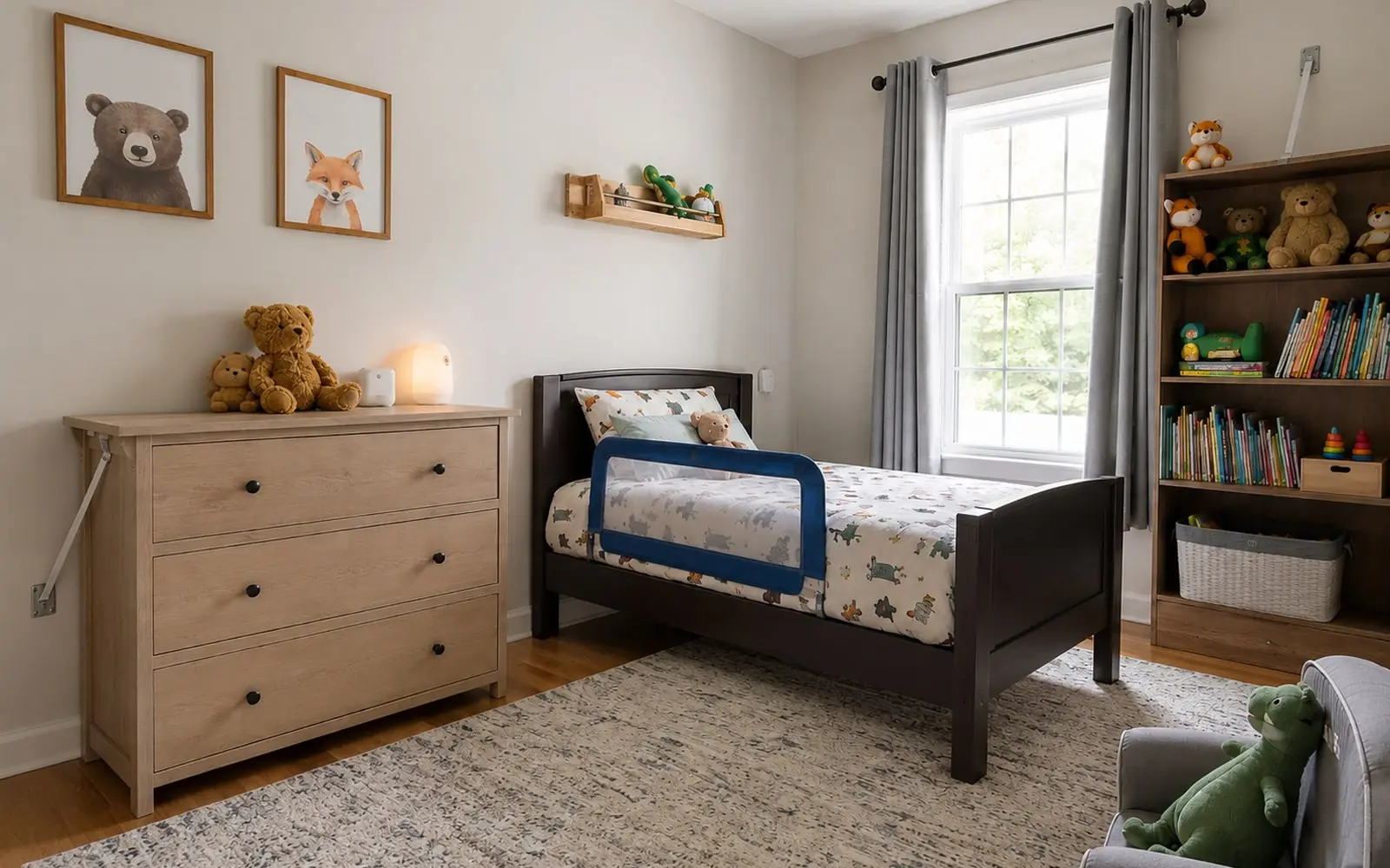 A toddler's bedroom with a low four-drawer dresser strapped to the wall, a twin bed with a guardrail, and a bookcase anchored beside the window.