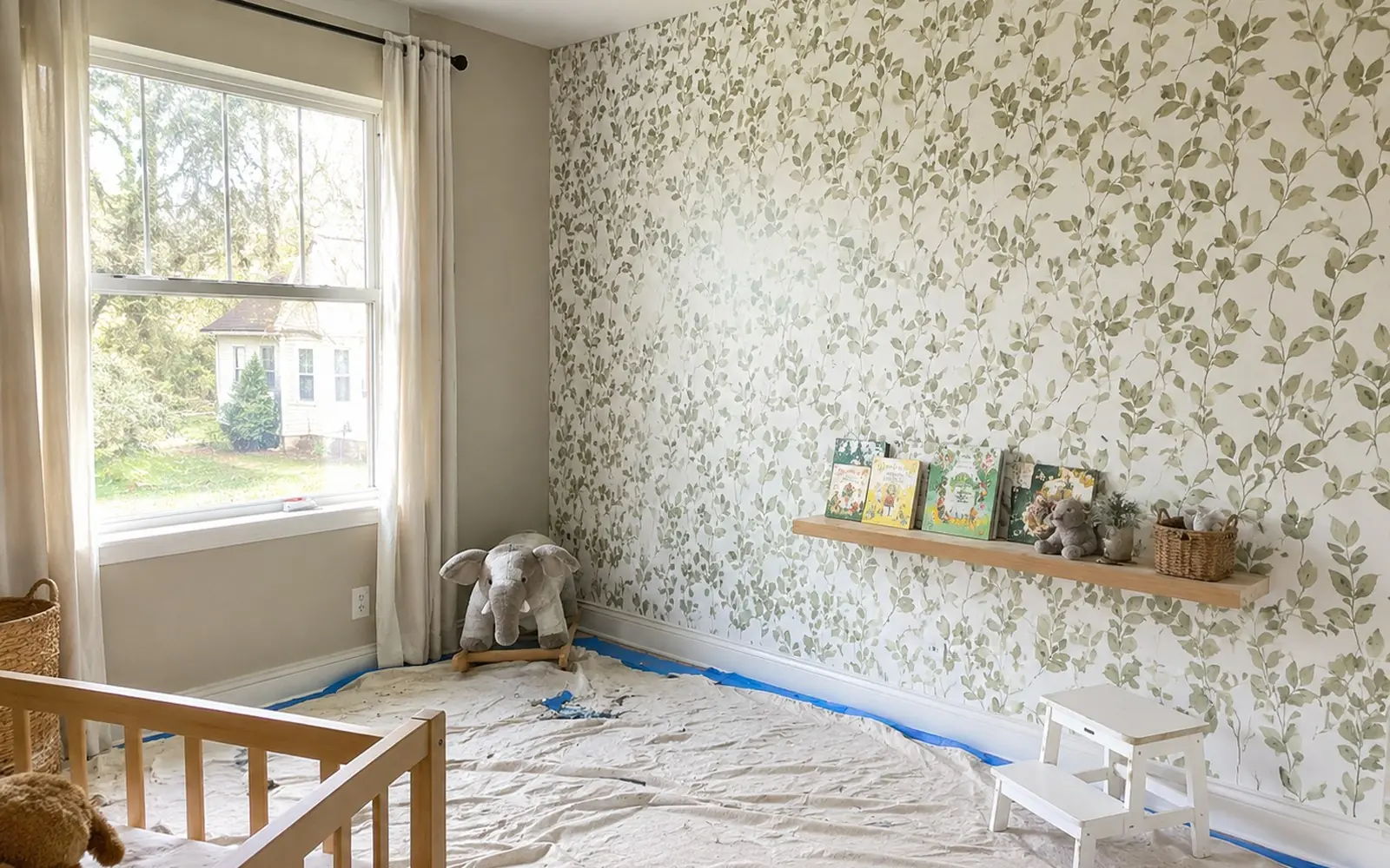 A child's bedroom mid-decorating, with one wall papered in a leaf pattern, a drop cloth taped down over the floor, and an anchored low shelf holding books.