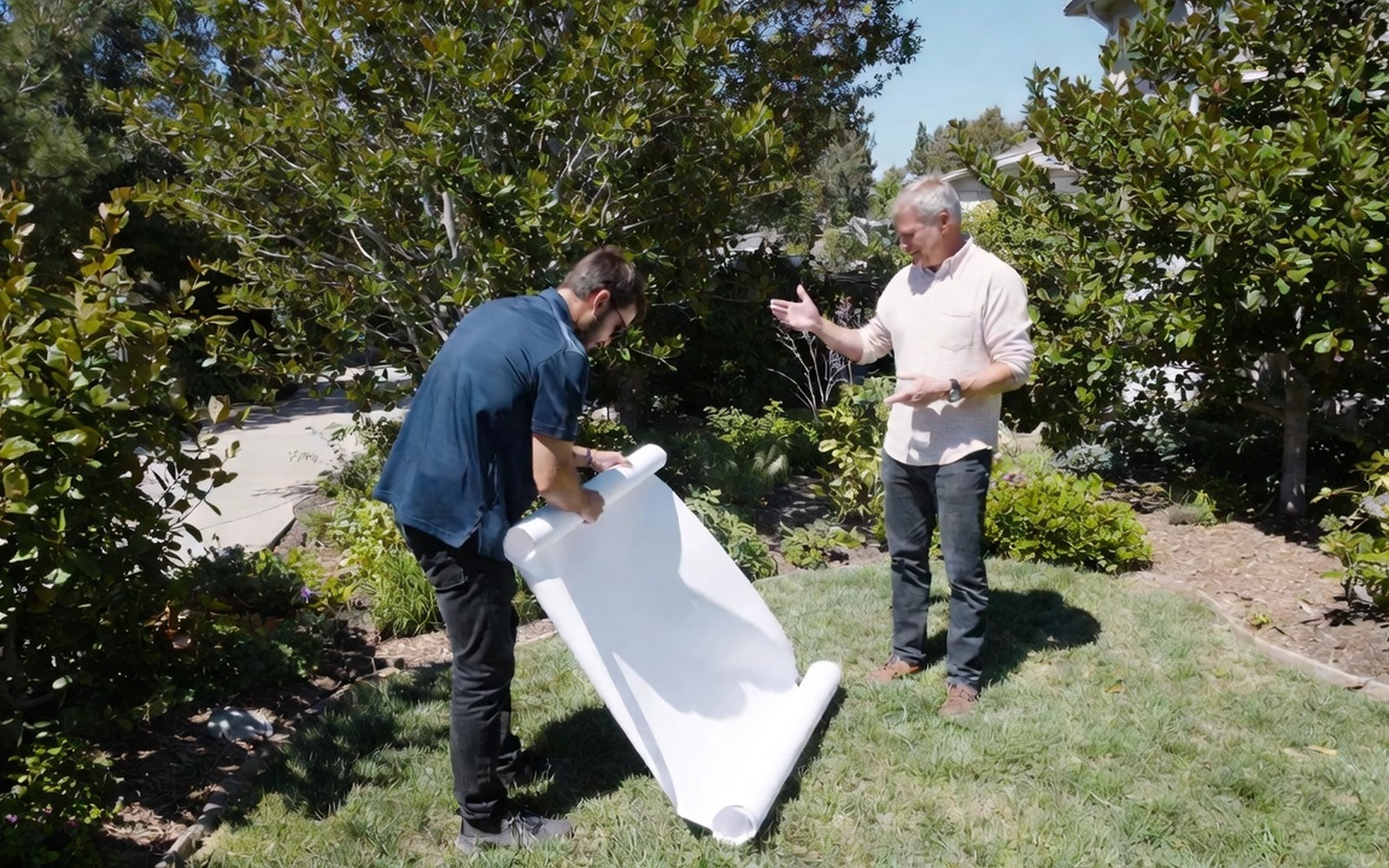 Designer unrolling an unreadable plan sheet in a yard with the homeowner, gesturing at beds