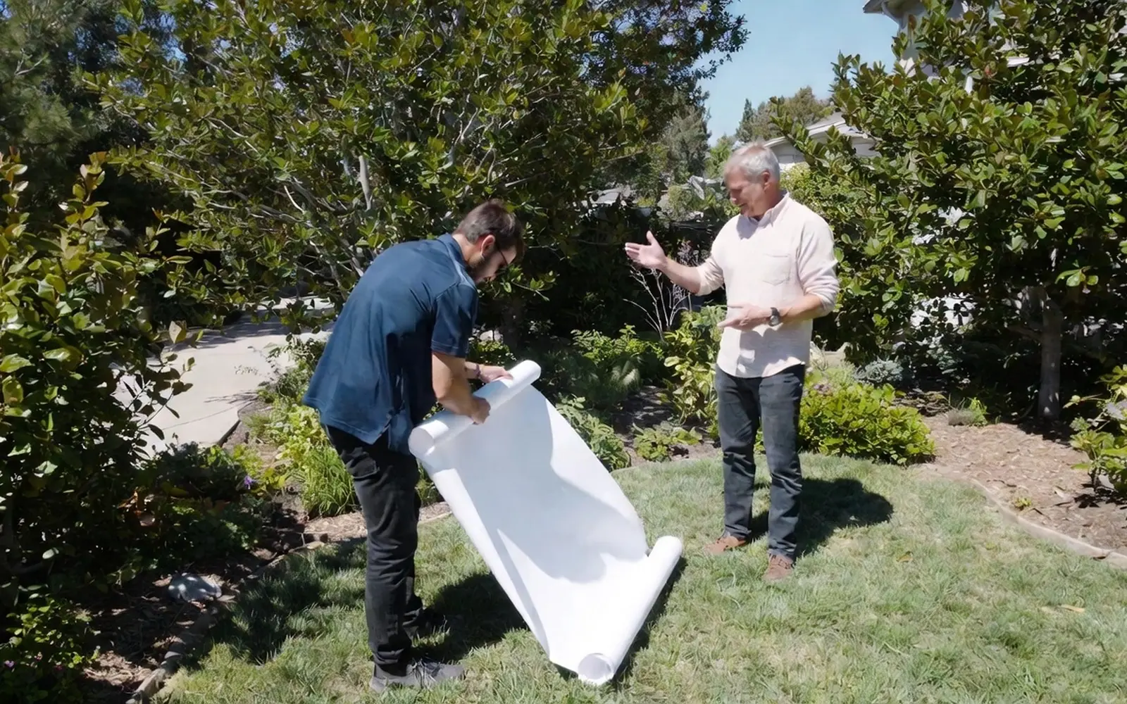 Designer unrolling an unreadable plan sheet in a yard with the homeowner, gesturing at beds