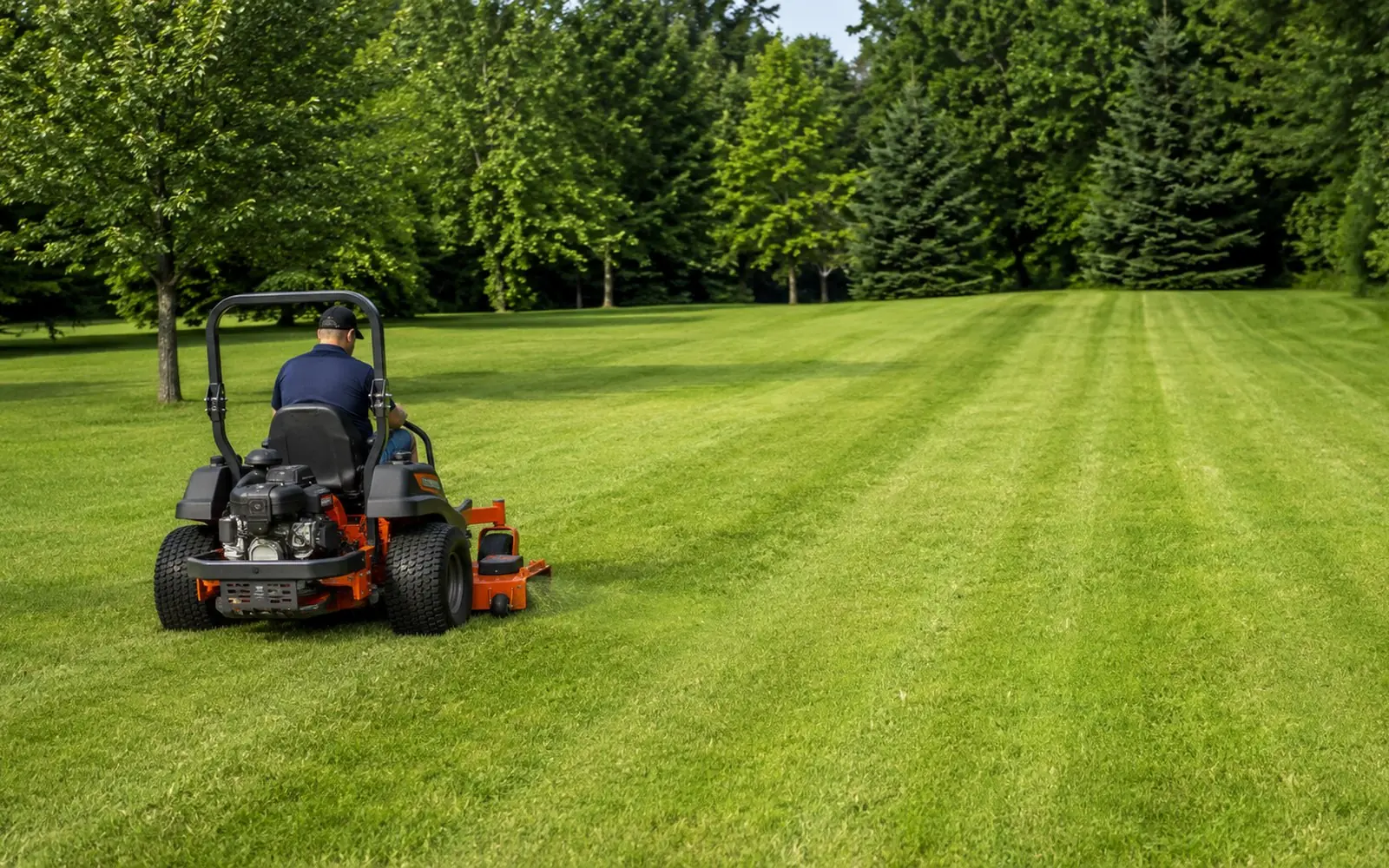A zero-turn riding mower cutting a wide stripe across a large open lawn bordered by mature trees, with cut lines visible behind it.