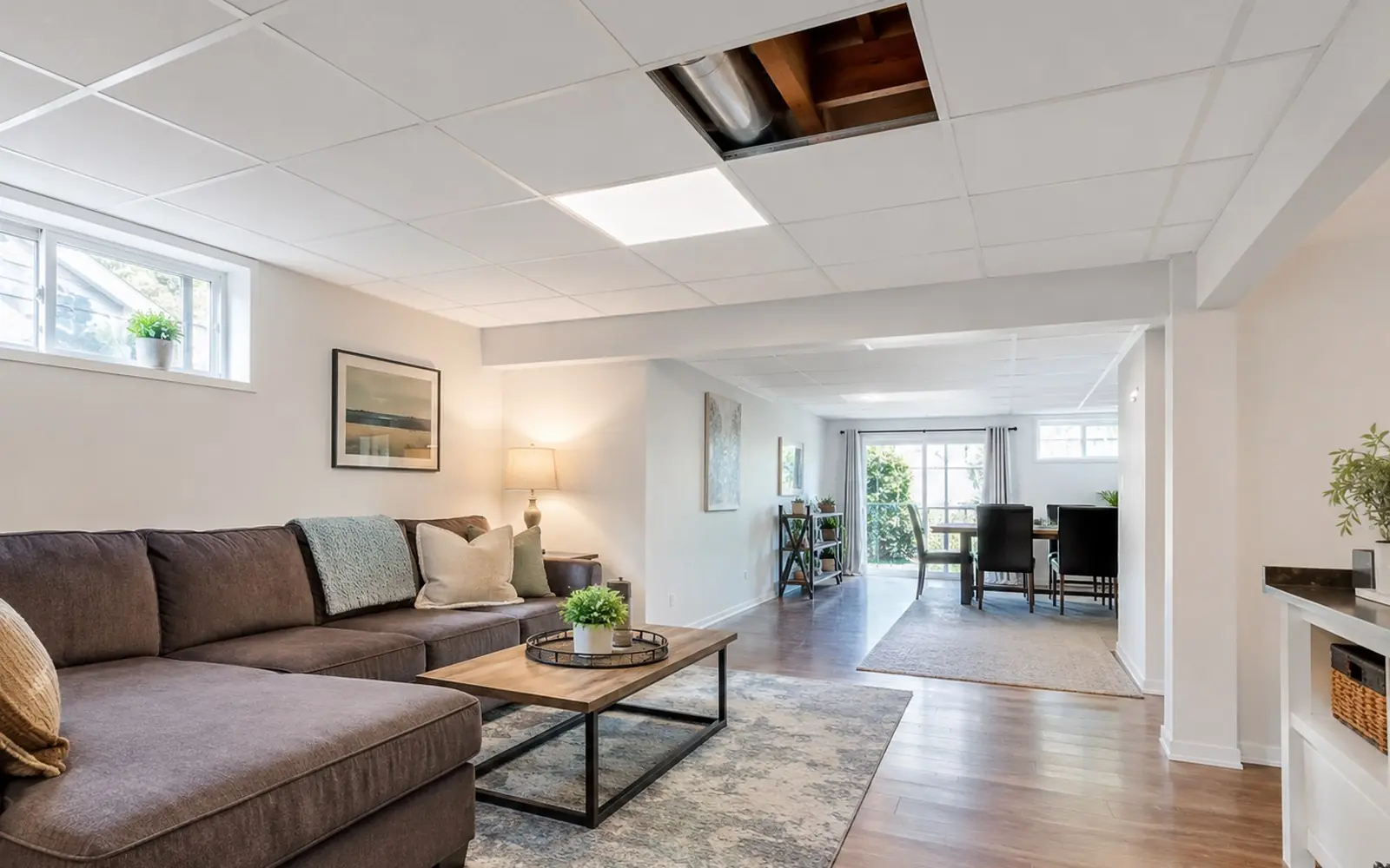 A basement room with a white suspended grid ceiling, one tile lifted out to show the joists and ductwork above, and a flat panel light seated in the grid.