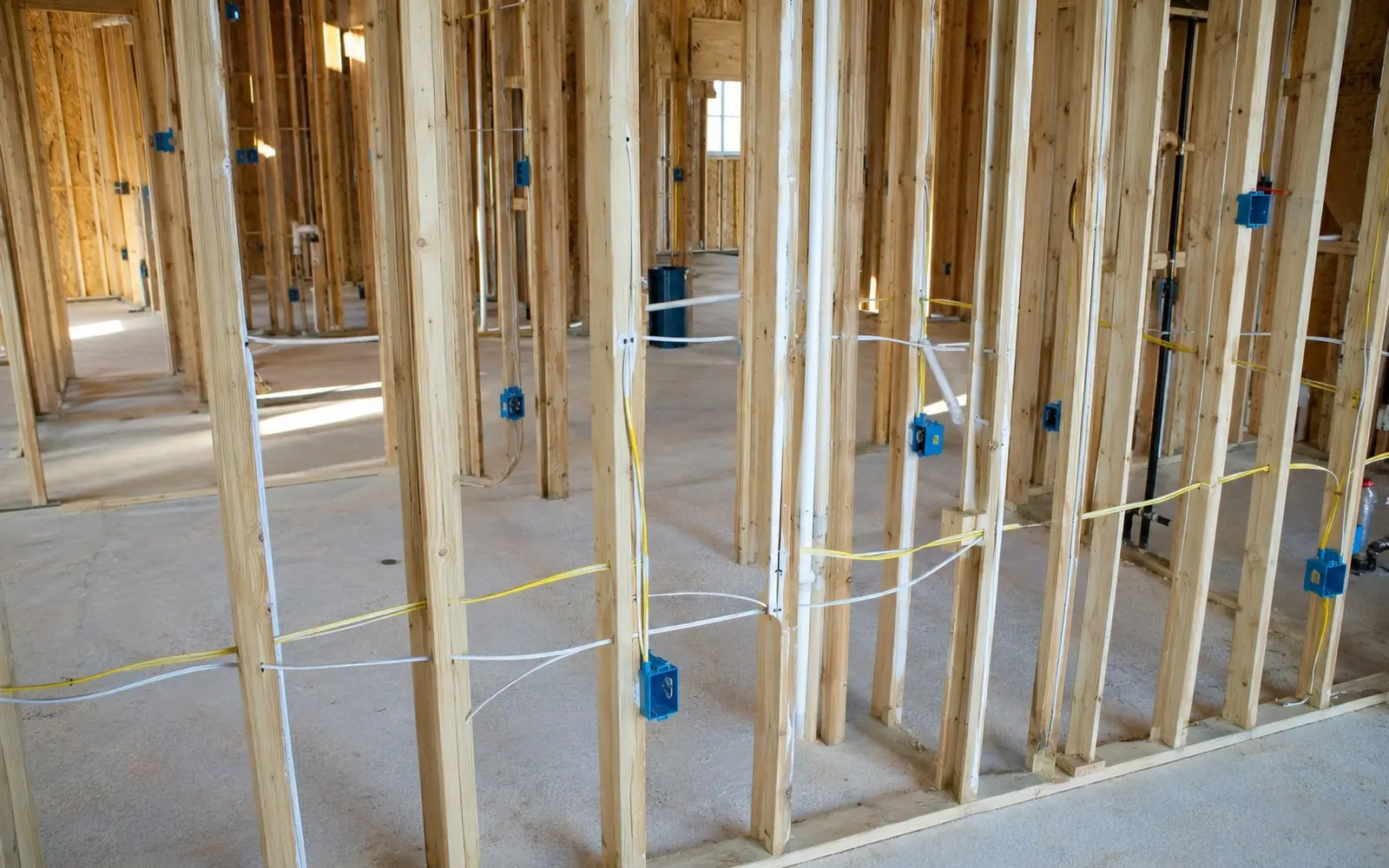 Open stud bays in a house under construction with yellow and white nonmetallic cable stapled to the framing and blue outlet boxes nailed in place.