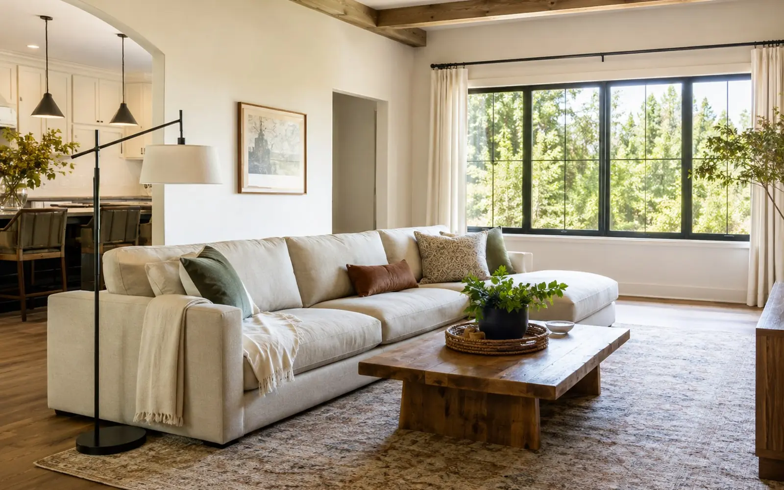 A sunlit family room with a fabric sectional facing a low wooden coffee table, a large area rug underneath, and a floor lamp beside the sofa arm.
