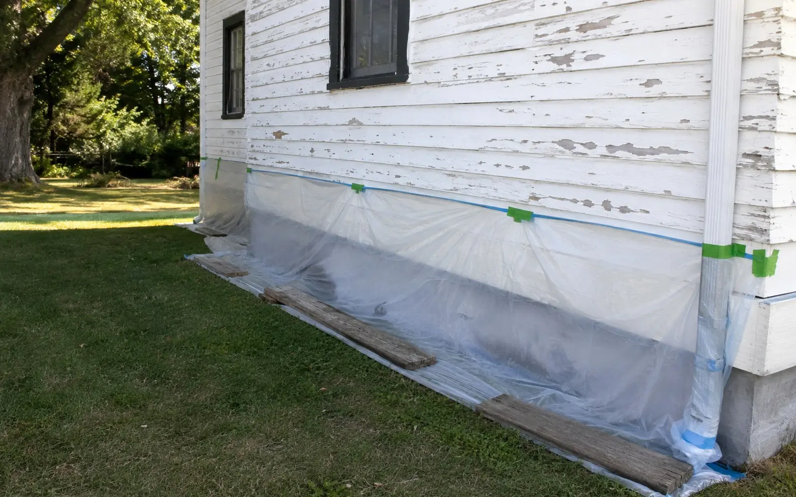 Plastic sheeting taped along the base of a weatherboard house with peeling white paint, held down with boards while an exterior wash is prepared.