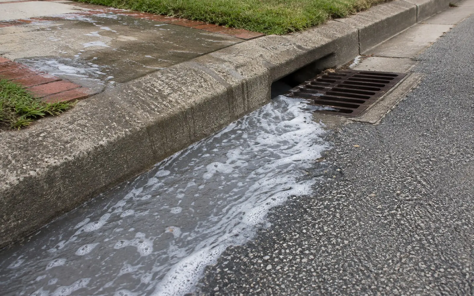 Grey soapy runoff from a driveway wash flowing along a concrete curb gutter toward an open storm drain grate.
